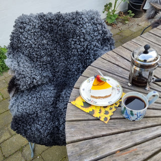 Gråt lammeskind med tætte krøller på en havestol ved et bord med kaffe og kage.
