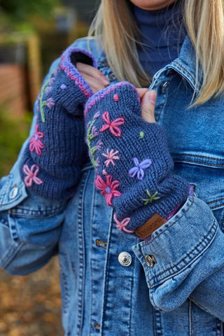 Blå pulsvarmere i uld med broderede blomster og lilla kanter, vist på hænderne.
