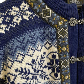 Den blå norske uldcardigans lyse mønster, bånd og tinhægter, vist tæt på.