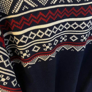Røde, hvide og gyldne mønsterbånd på den marineblå merinouldsweater, vist tæt på.